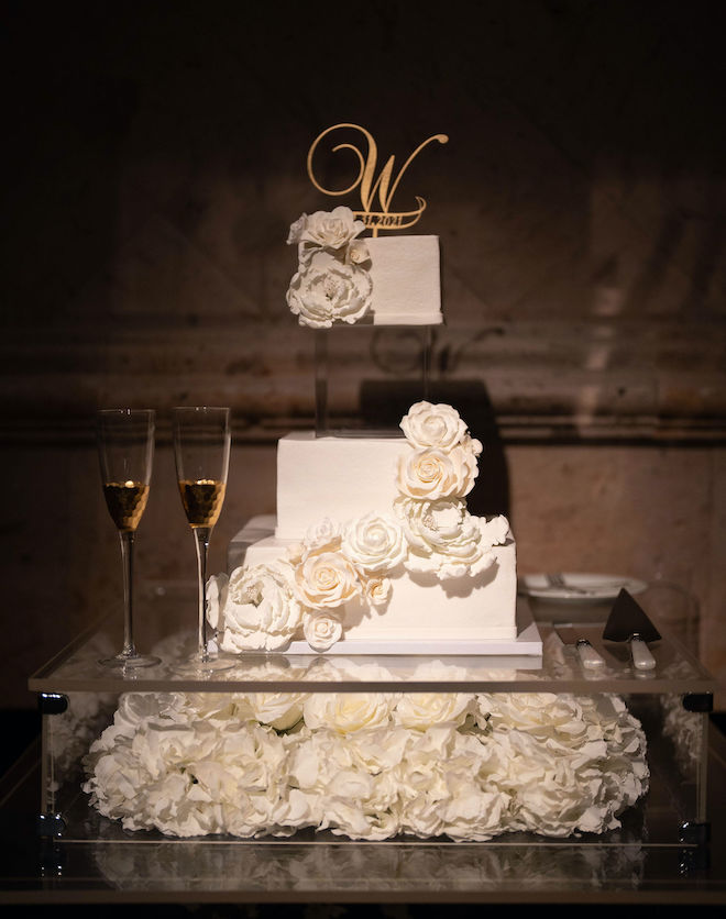 A white wedding cake decorated with white roses sits on a clear cake stand. 