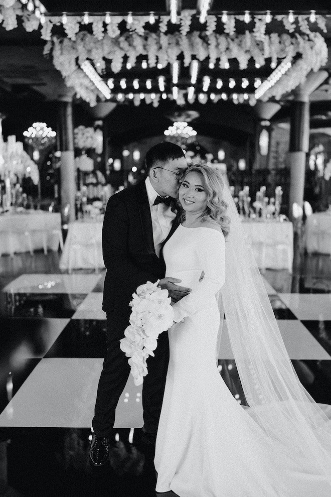 Groom kisses a bride wearing a long sleeve Ines Di Santo gown in a ballroom decorated for a wedding reception at The Astorian in Houston, TX.