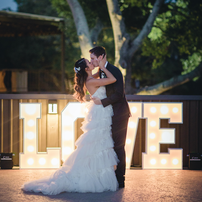 Bride and groom kiss in front of a lit sign that reads, "love" 