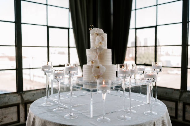 White wedding cake garnished with white lillies surrounded by clear candle holders. 