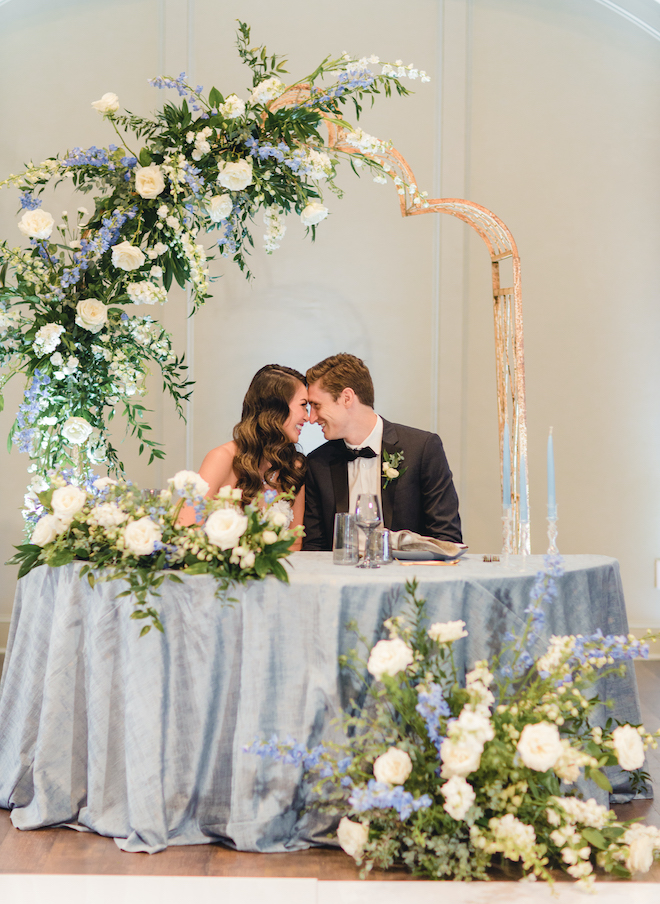 Bride and groom smile while touching foreheads at a sweetheart table surrounded by florals, greenery and a small arbor at a wedding reception at Pine FOrest Country CLub. 