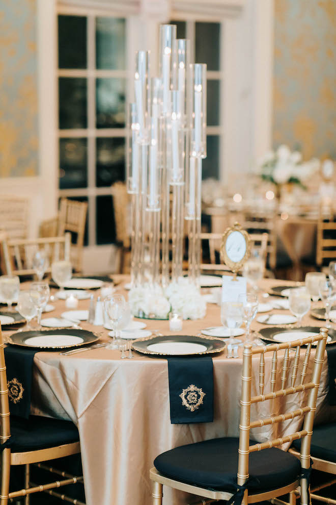 round banquet table set with gold tablecloth, gold and black placesettings and lit tapered candles in tall glass candleholders at a wedding reception at the Junior League in Houston, TX.