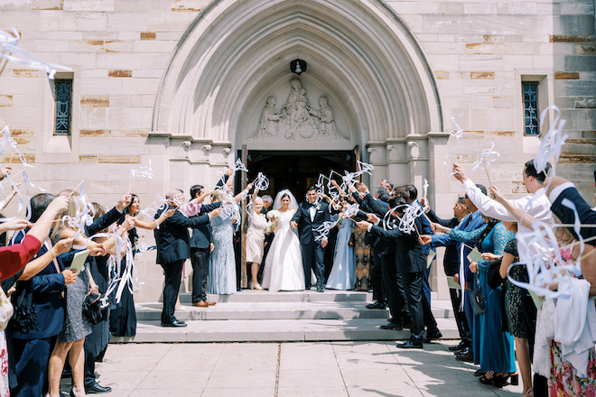 The bride and groom leaving the church while the guests wave streamers.