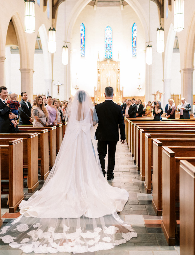 The bride wearing a satin Sareh Nouri dress with a bow on the back and a lace veil, walking down the aisle with her father.