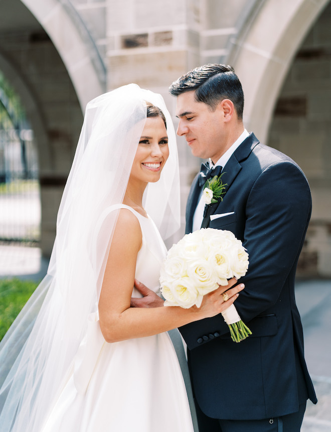 The groom looking at the bride as she smiles in her white satin gown and veil.