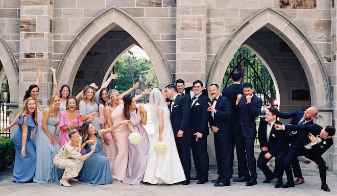 The bride and groom kissing while the wedding party points at them.