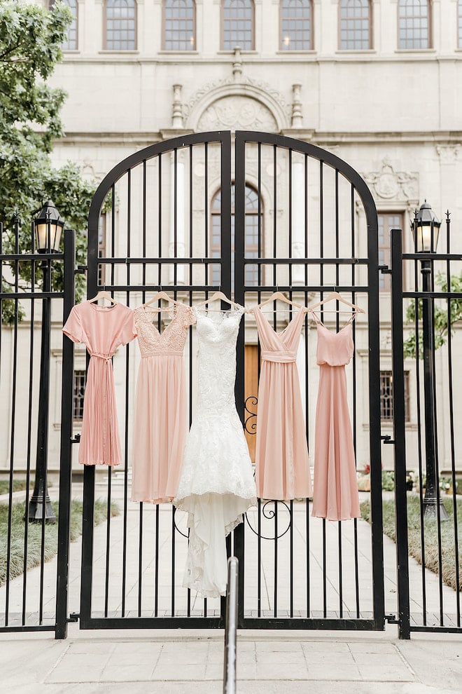 The bridal gown from Parvani Vida and the blush bridesmaids dresses hanging on the gate outside Julia Ideson library.