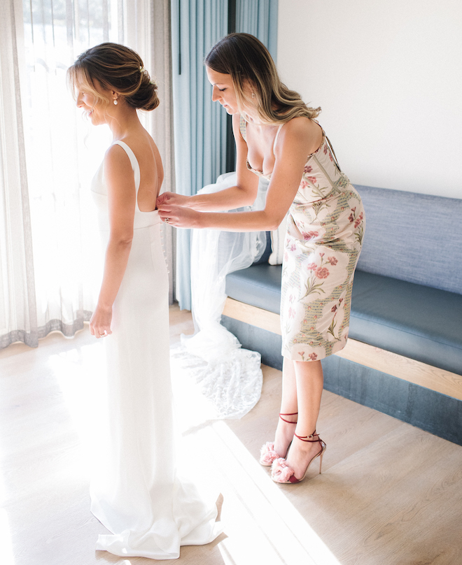 A bridesmaid buttoning the back of the bride's wedding gown before the alfresco ceremony.