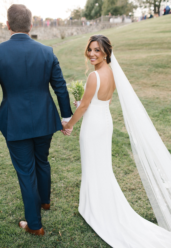 The bride looking back as she and her husband walk holding hands at their outdoor autumn wedding.