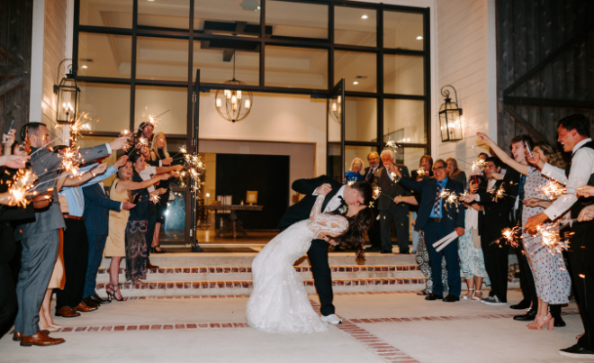 The bride and groom share a kiss as they exit their pastel-colored wedding while family and friends cheer with sparklers. 