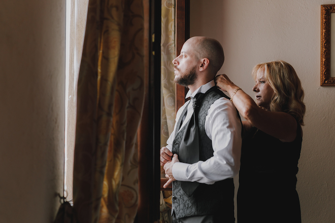 A woman helping the groom adjust his suit as he looks out the window.