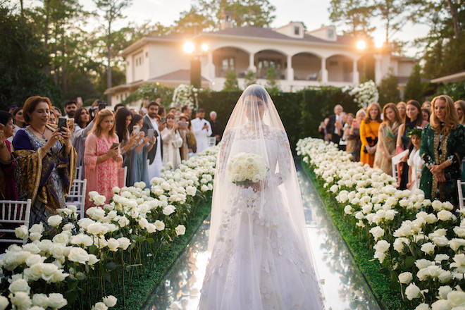 The bride walking down the aisle at while greenery and white roses adorn this backyard garden ceremony.