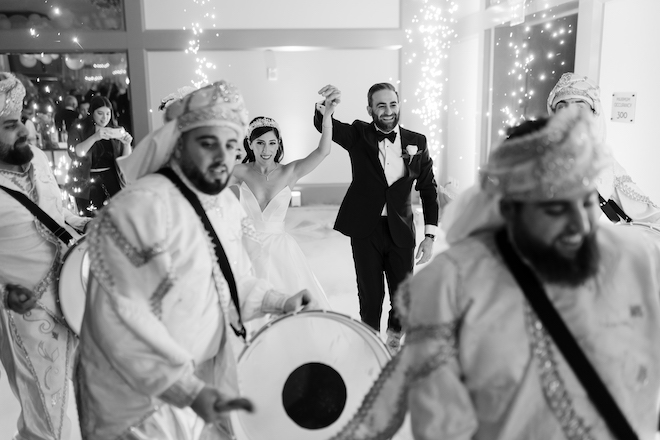 The bride and groom holding hands walking behind men playing drums and sparklers going off.