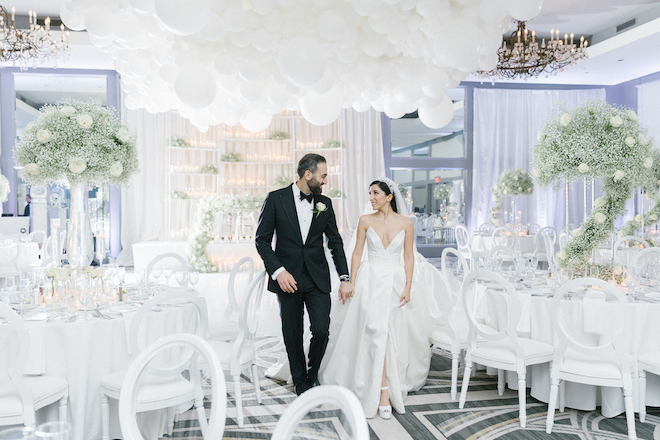 The bride and groom holding hands in the all-white decorated ballroom.