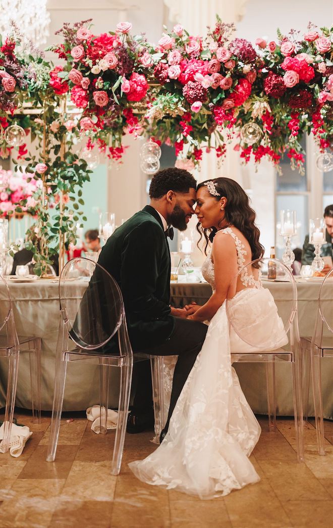 A groom and bride sit face to face in acrylic ghost chairs at a wedding party table set with florals, hanging orbs and lit candles. 