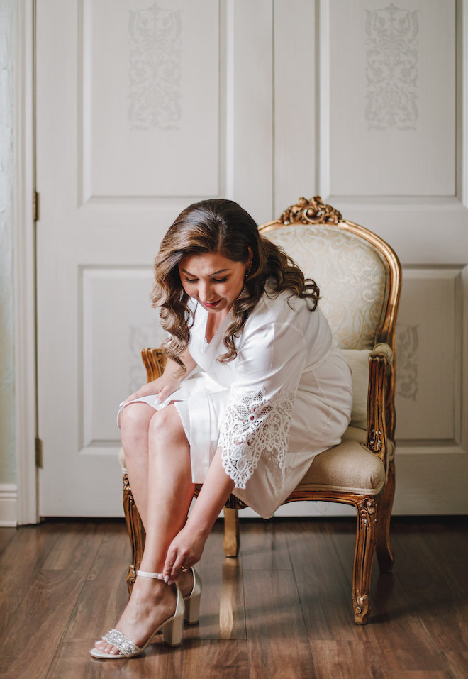 The bride wearing a white robe putting on heels with a sparkle strap. 