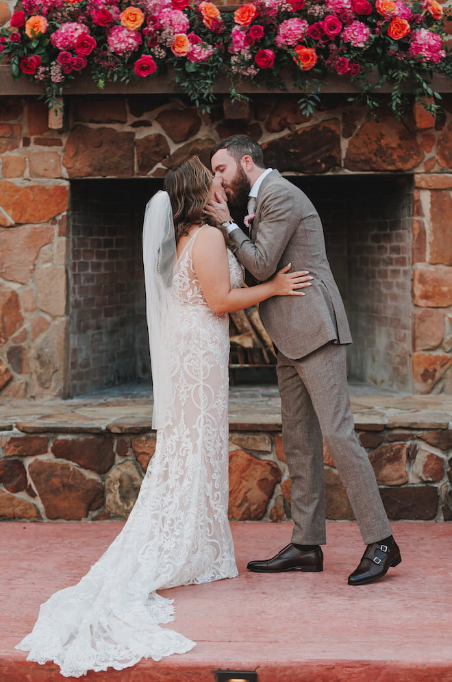 The bride and groom kissing at their alfresco ceremony with pink and orange florals above them decorating the altar. 