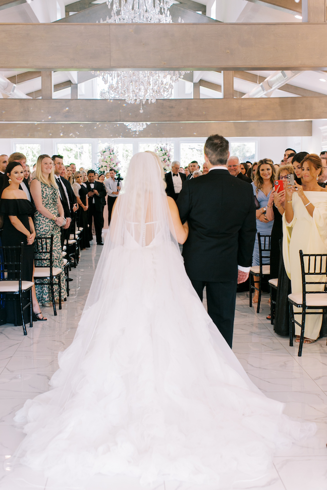 The backs of the bride and her father walking down the aisle while guests smile at them and take photos. 