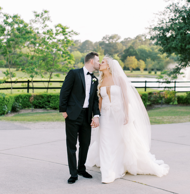 The bride and groom outside holding hands kissing in front of a green field and creek. 