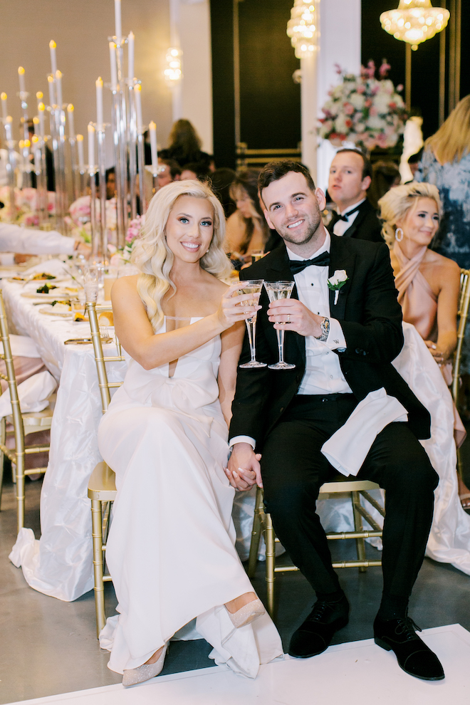 The bride and groom clinking champagne glasses sitting at the reception table.