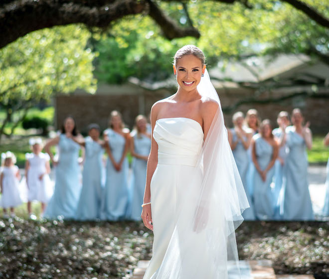 The bride smiling while her bridesmaids in bright blue dresses react to seeing her for the first time.