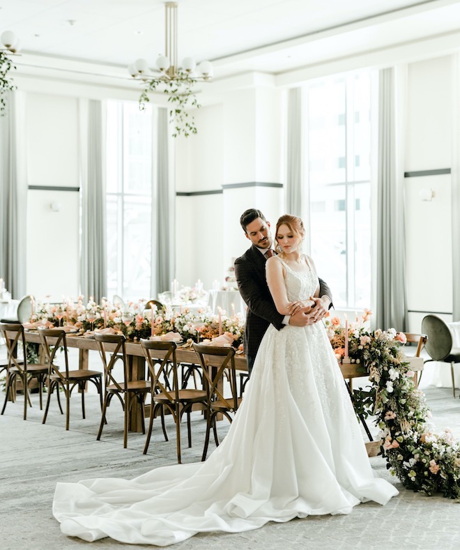 A groom hugging the bride in front of a dining table in the ballroom of The Laura Hotel.