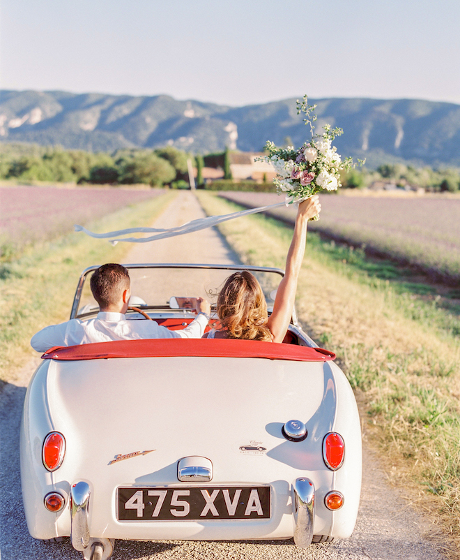 The bride and groom driving away in a vintage car as the bride holds up her bouquet.