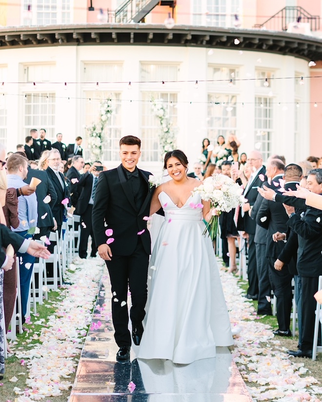 A bride and groom walking down the aisle in front of Centennial Greens lined with flower petals. Guests are throwing flower petals as the bride and groom walk. 