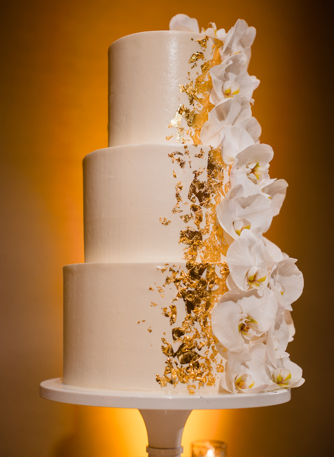 A three tier white wedding cake with gold flakes and white flowers.