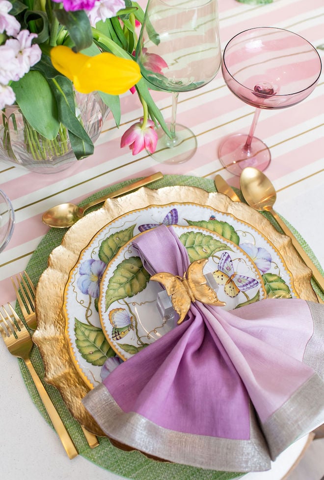 A colorful tabletop with purple and green china and linens, gold flatware and pink and green colored-glasses.