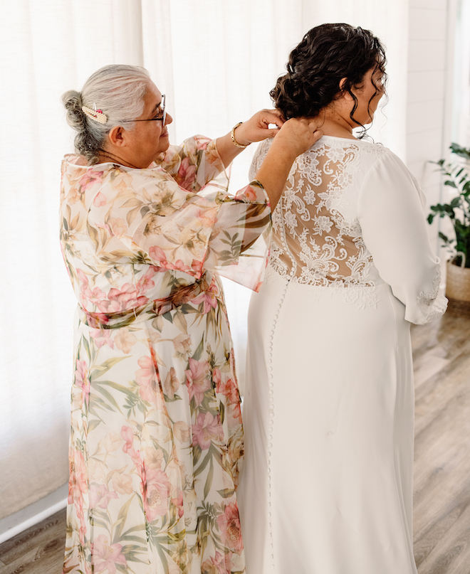 The bride's mother in a floral long dress buttoning the back of the bride's wedding gown.