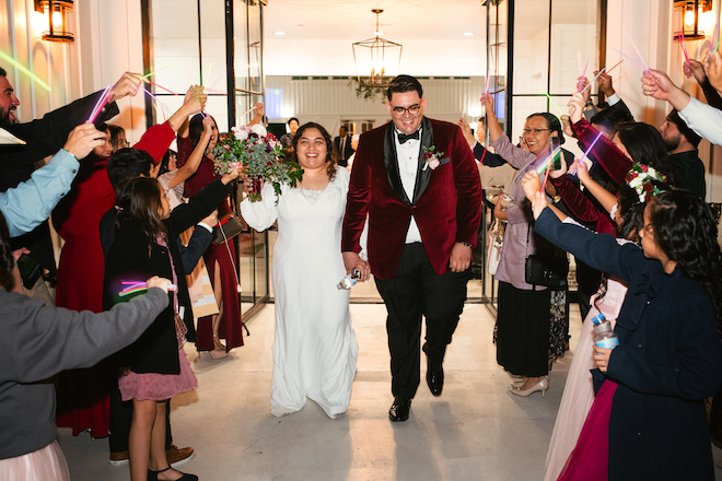 The bride and groom holding hands and smiling during their sendoff with guests waving glow sticks on either side of them. 