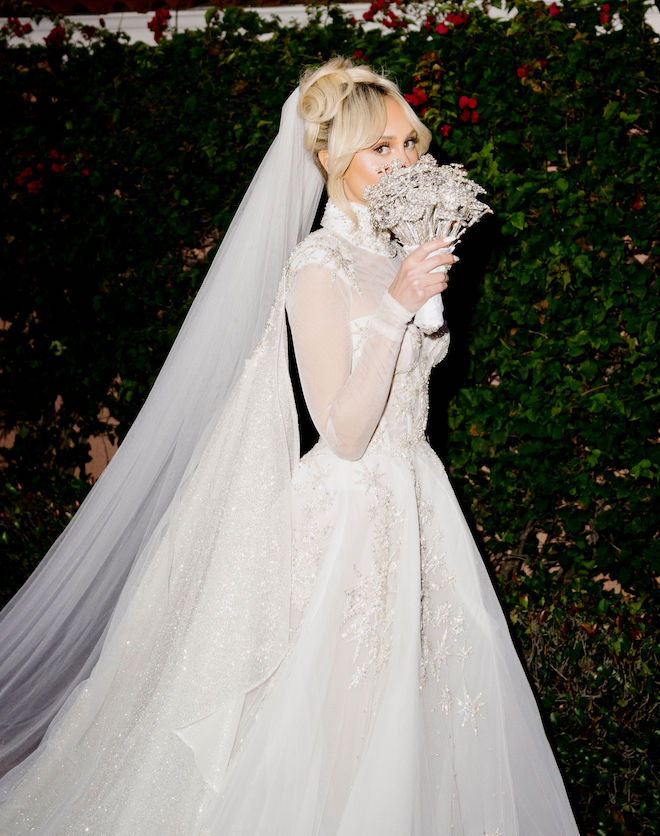 A bride wearing her wedding gown and veil holding up her all-crystal bouquet to her face.