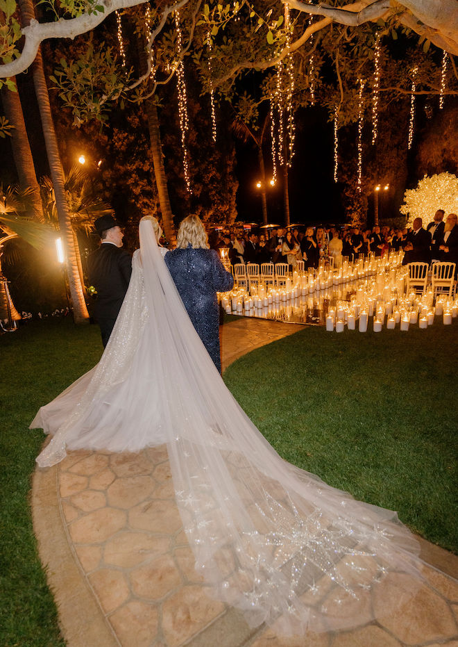 The bride and a man and woman on either side of her walking toward the candle-lit aisle.