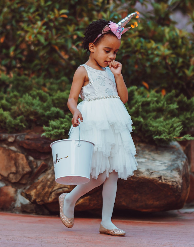 The flower girl wearing a white dress with a unicorn headband and holding a white bucket with her finger in her mouth. 