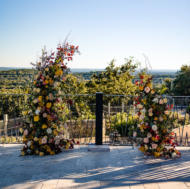 A floral arrangement with peach, gold, red flowers with greenery in a broken arch as the ceremony site. 