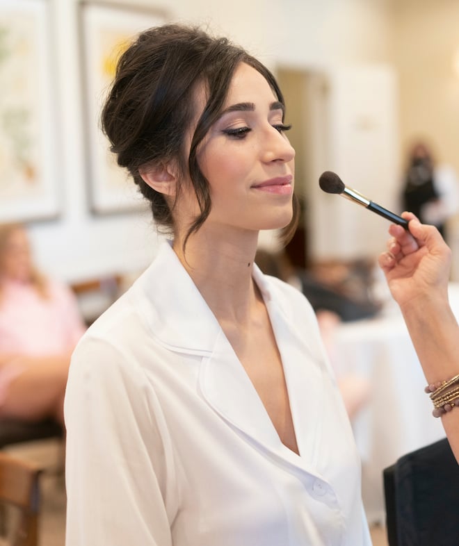 A bride with a formal updo has makeup applied on the morning of her wedding in Houston