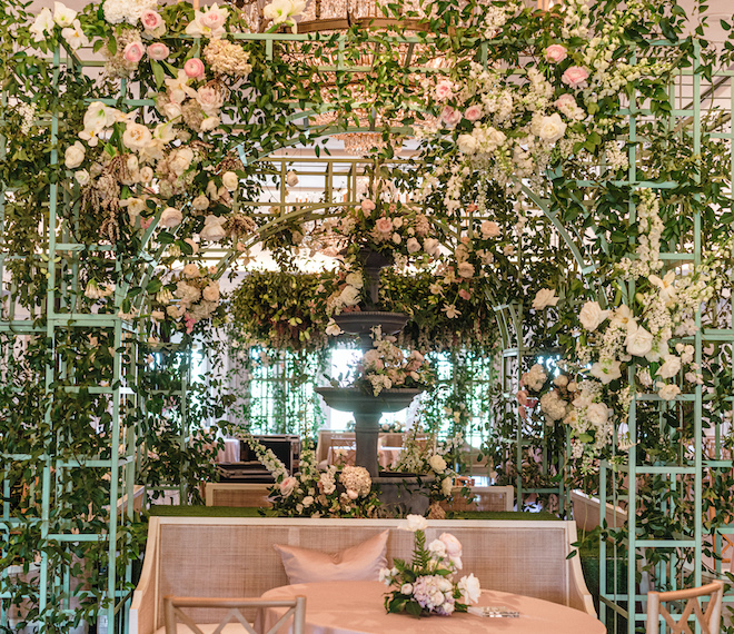 French inspired florals covering the French green treillage and a fountain in the center. A pink couch sits in front of it. 