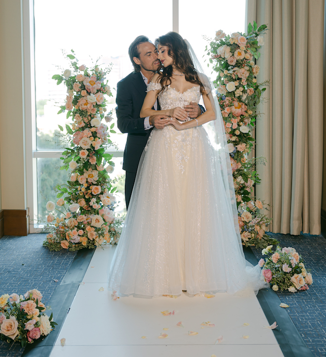 The groom hugging the bride from behind in front of the pink-floral altar with floor-to-ceiling windows behind them in the ballroom of The Westin Houston Memorial City.