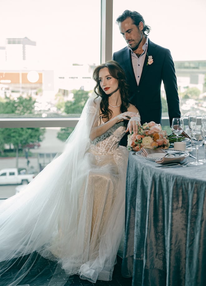 The bride sitting at the reception table with the groom standing behind looking down at her with floor-to-ceiling windows behind them in the ballroom of The Westin Houston Memorial City.