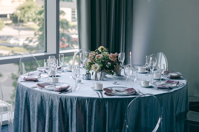 A circular reception table with a blue tablecloth, glass and white dinnerware and pink napkins in front of a window in the ballroom of The Westin Houston Memorial City.