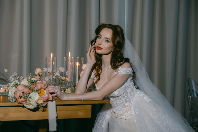 The bride resting her hands on the reception table while touching a pink bouquet of flowers. Pink and white candles glow behind her. 