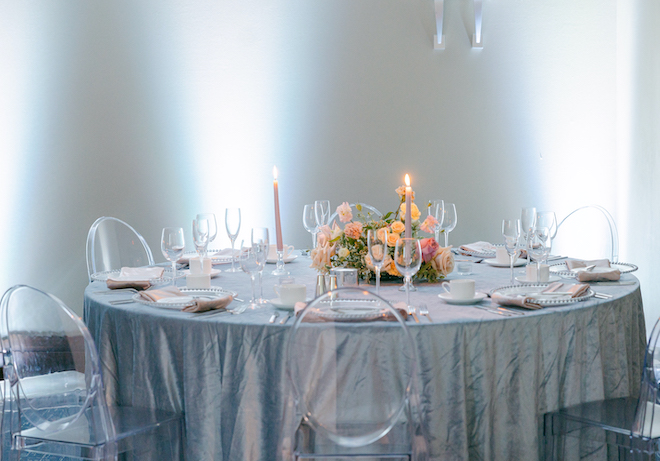 A circular reception table in a blue tablecloth with pink pillar candles and a pink and green floral arrangement as a centerpiece. 