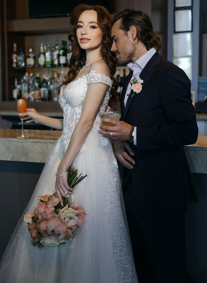 The groom standing behind the bride at the bar both holding cocktails. 