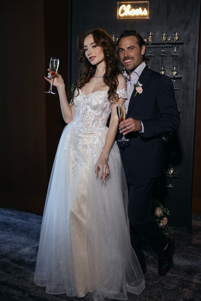 The bride and groom smiling holding glasses of champagne in front of a black champagne wall with a lit-up "Cheers" sign.