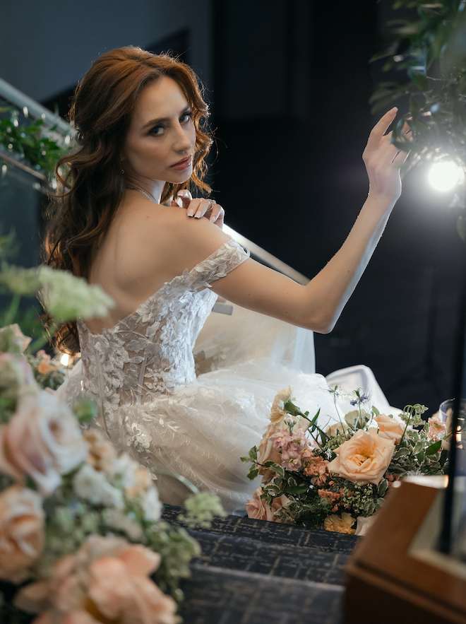 The bride looking behind while sitting on the staircase covered with pink-hued florals. 