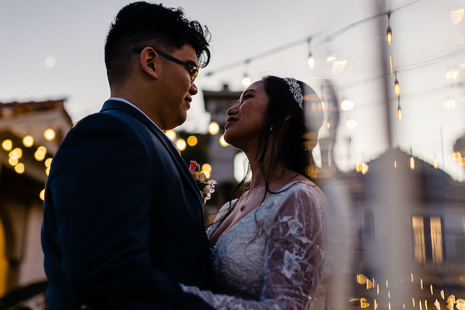 The bride and groom smiling at each other under twinkling lights. 