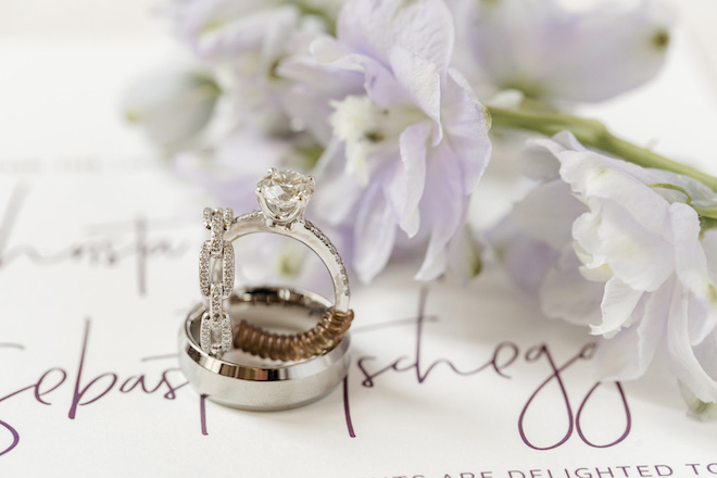 Two diamond rings sitting on top of a silver wedding band with a white invitation with purple font and a purple flower lay next to it. 