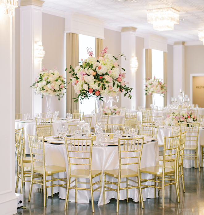 White round tables with gold chairs and large white and pink floral centerpieces. 