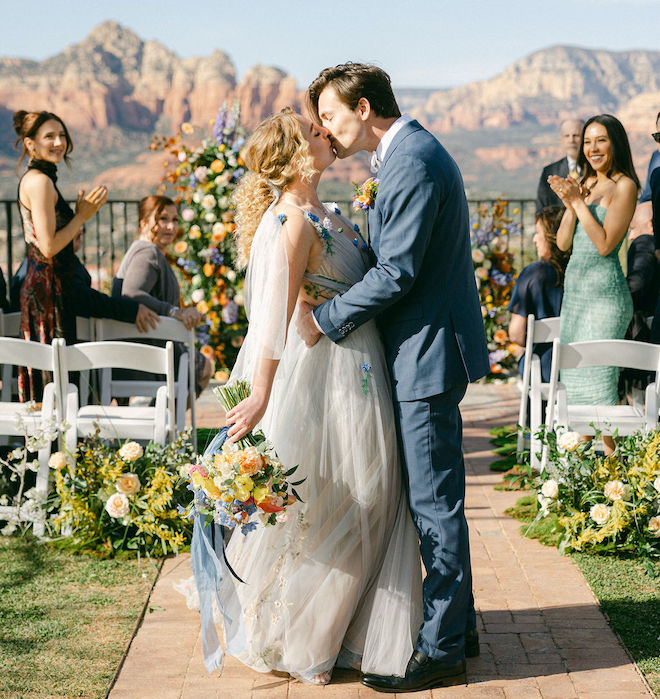 The bride and groom kissing while their guests clap for them after their vibrant wedding ceremony. 